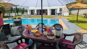 An outdoor dining area with a pool in the background, surrounded by a modern house and greenery. - Villa Aronora Private Pool & BBQ (Haddada)