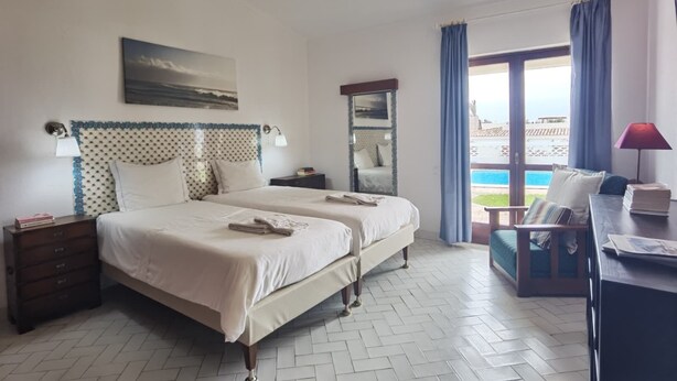 A hotel room with a large bed, two bedside tables, a desk with a lamp, a view of the sea through a large window, and a balcony. - TP Maria Luisa dos Olhos Villa - Private Pool and Beach (Olhos de Água)