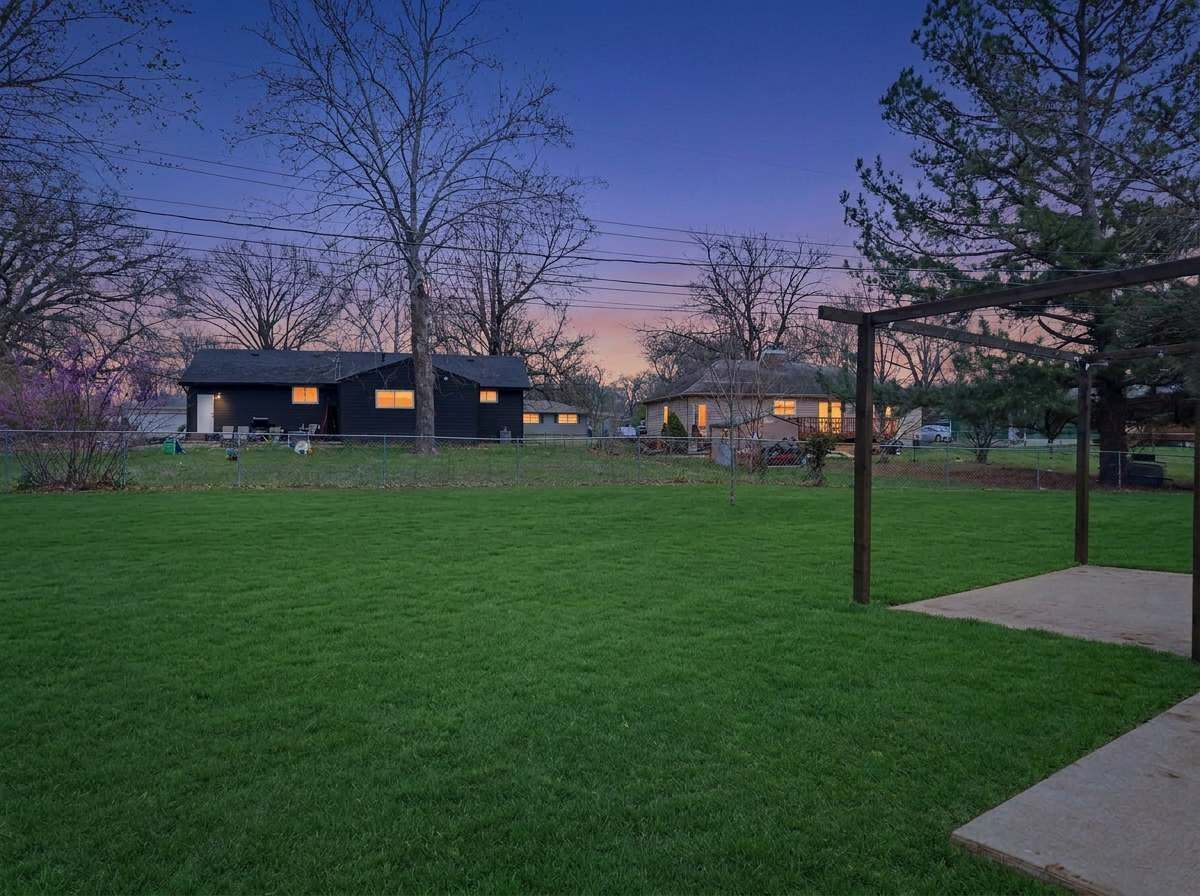 Un quartier résidentiel avec une pelouse verdoyante, une pergola en bois et des maisons dont les fenêtres sont illuminées.