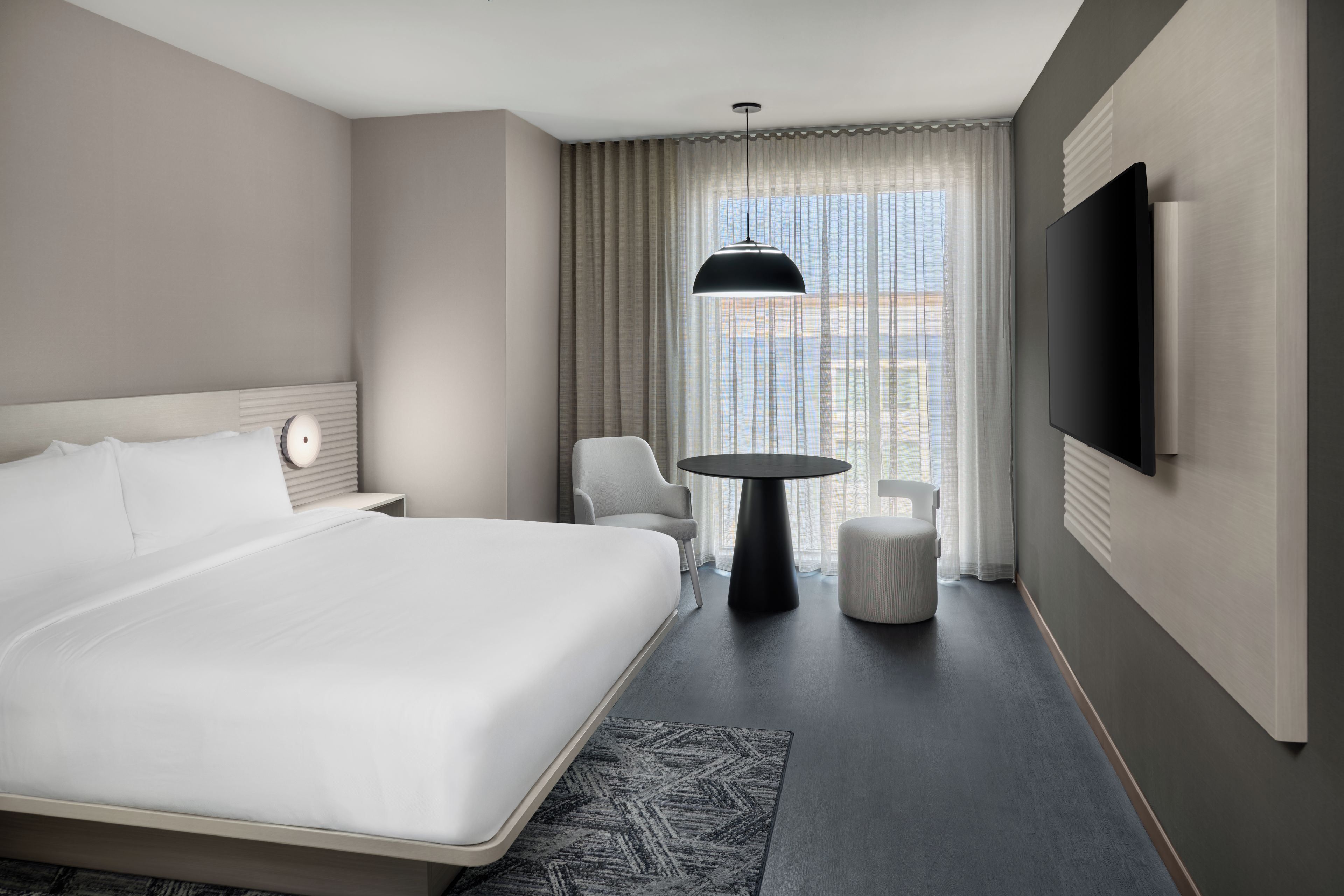 A hotel room with a large bed, a round table, a chair, a TV, and a window with curtains.