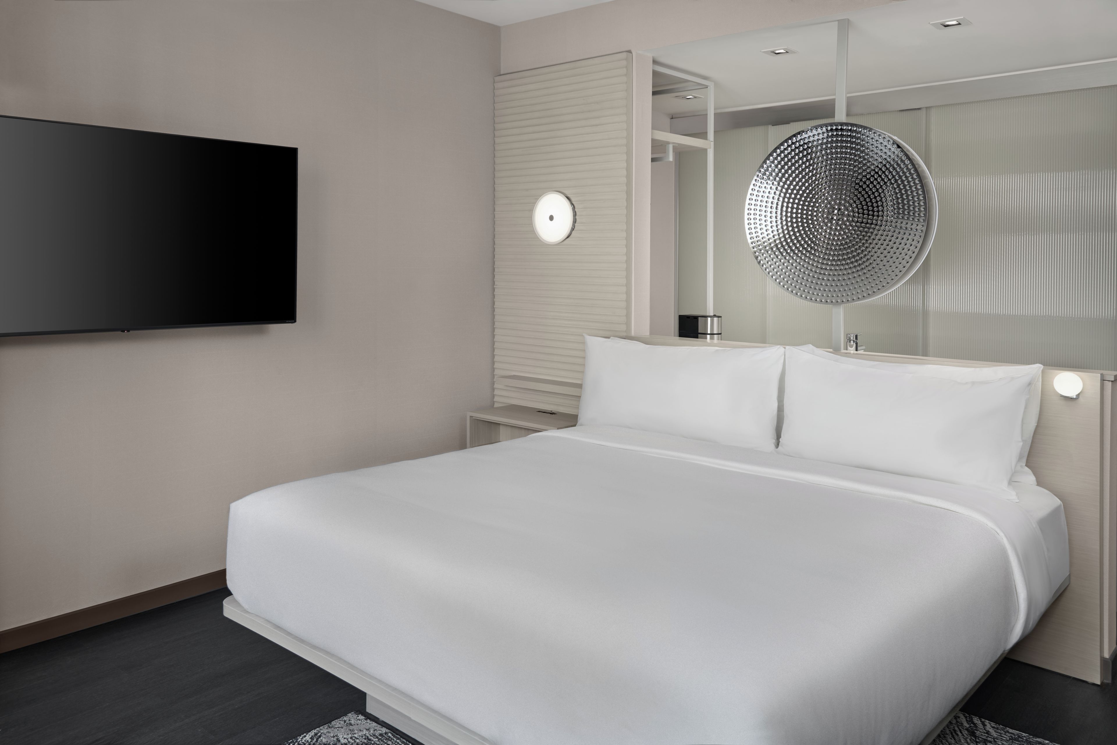 Habitación de hotel moderna con una cama grande, un televisor de pantalla plana montado en la pared, un espejo redondo con un marco decorativo y una mesita de noche pequeña.