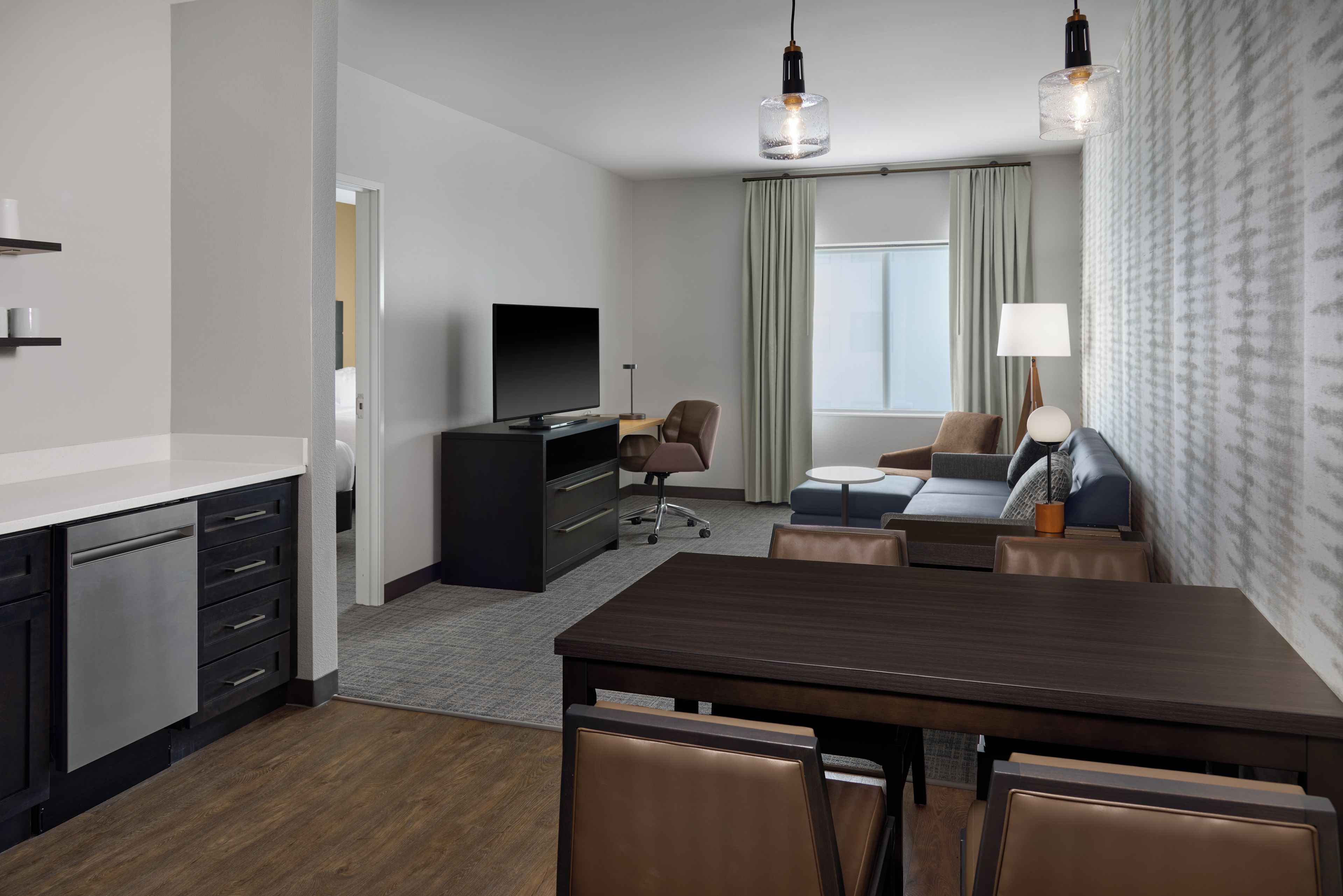 A modern hotel room with a dining area, a flat-screen TV, a sofa, a lamp, and a window with curtains.