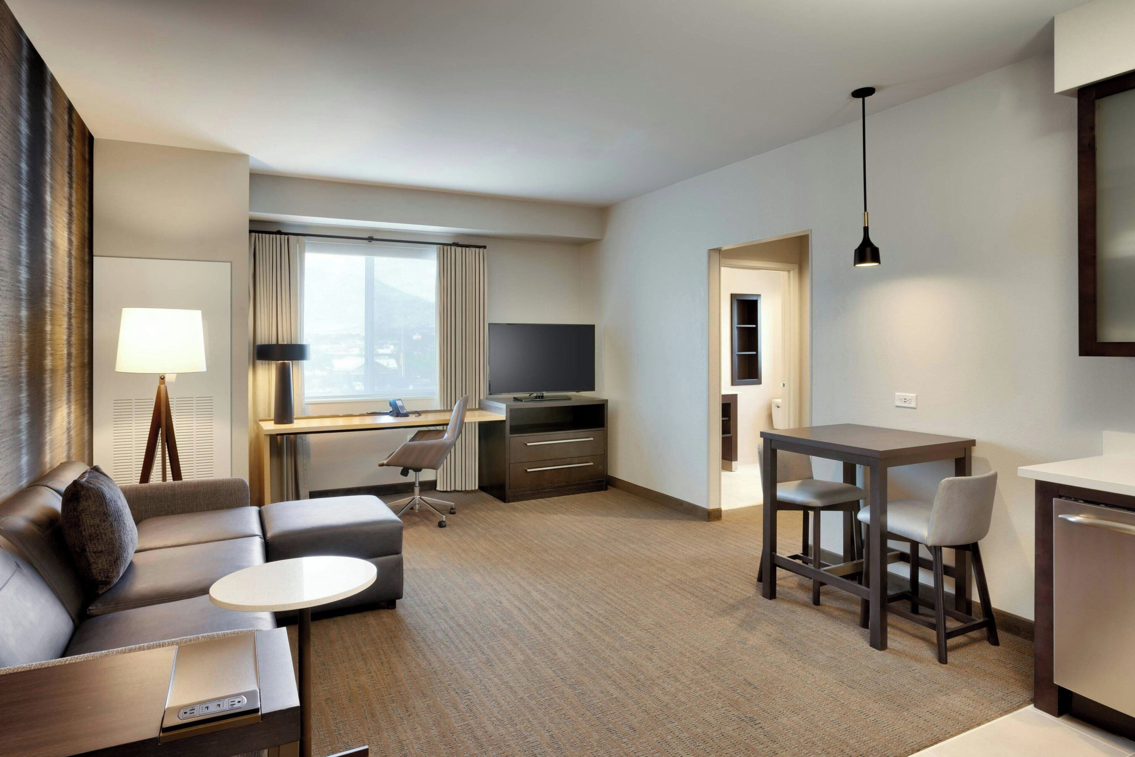 Habitación de hotel moderna con sofá, una pequeña mesa redonda, escritorio con computadora, televisor y comedor con sillas.
