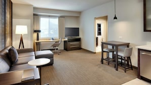 Habitación de hotel moderna con sofá, una pequeña mesa redonda, escritorio con computadora, televisor y comedor con sillas.