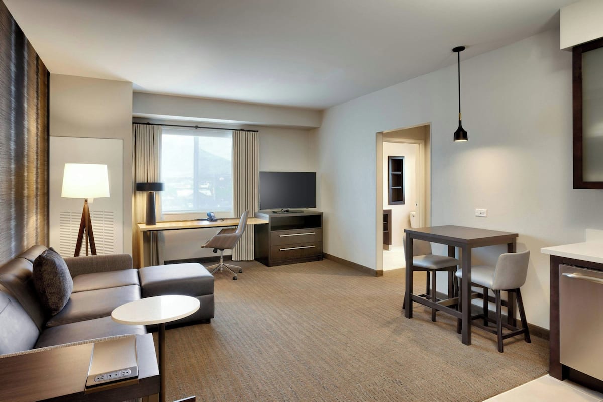 Habitación de hotel moderna con sofá, una pequeña mesa redonda, escritorio con computadora, televisor y comedor con sillas.