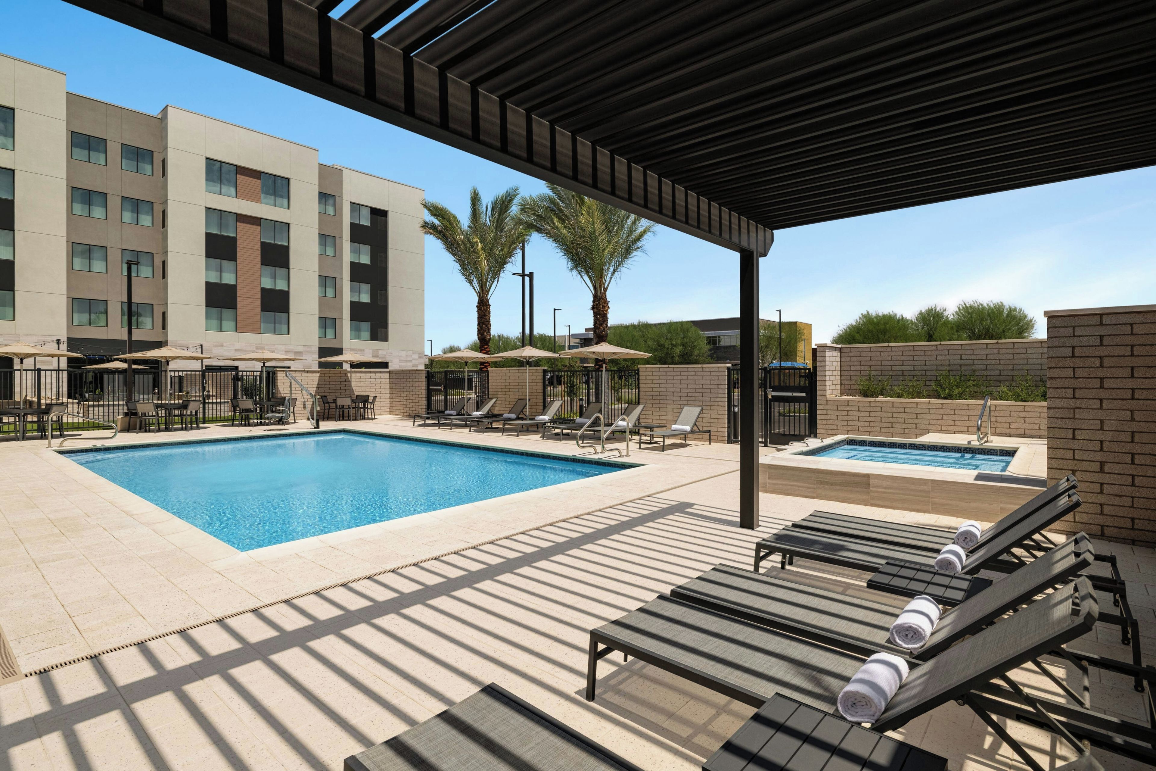A pool area with lounge chairs and umbrellas, a separate hot tub, and a building with multiple windows in the background.