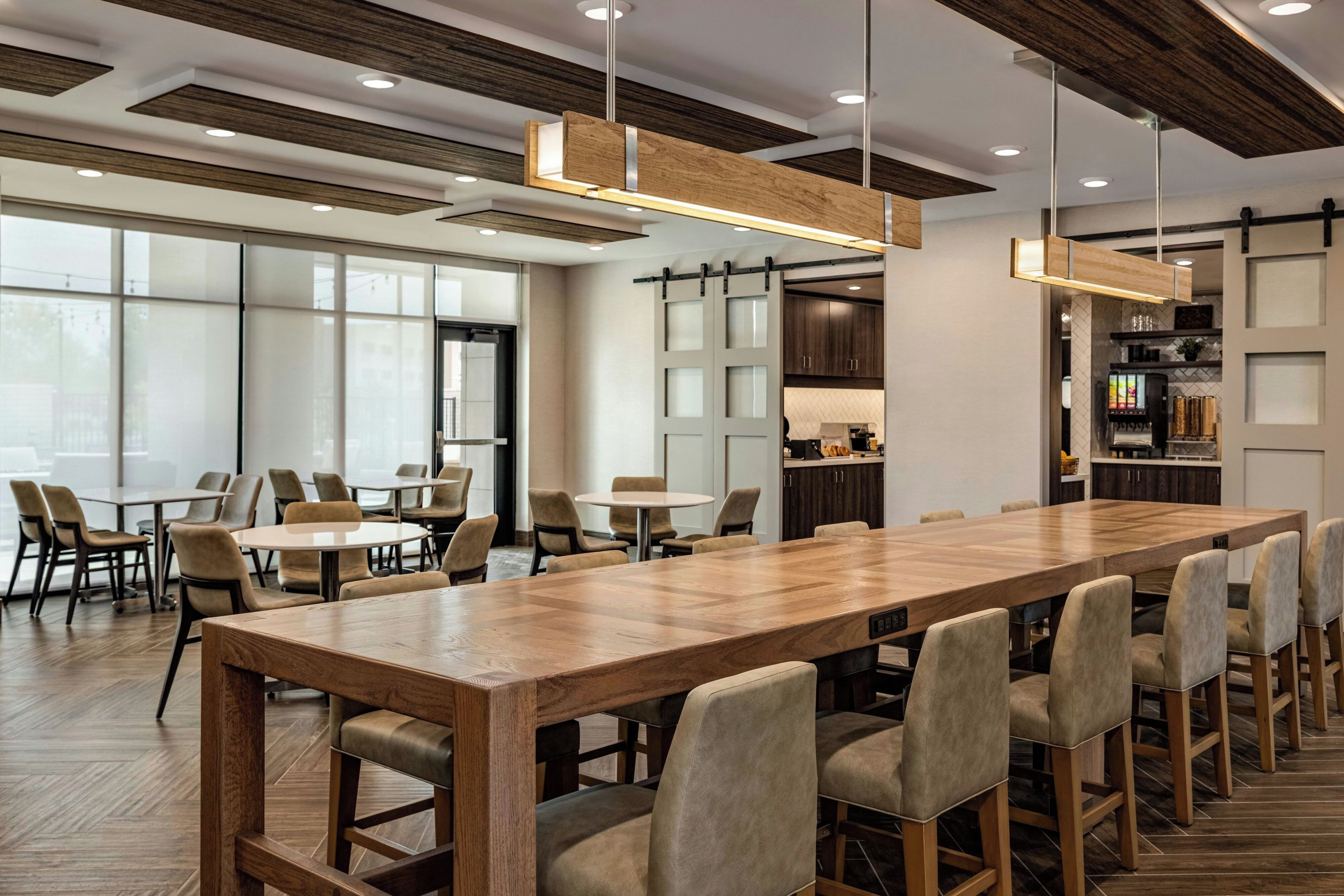 A spacious dining area with a long wooden table and multiple chairs, a kitchen area with dark wooden cabinets, and large windows allowing natural light.