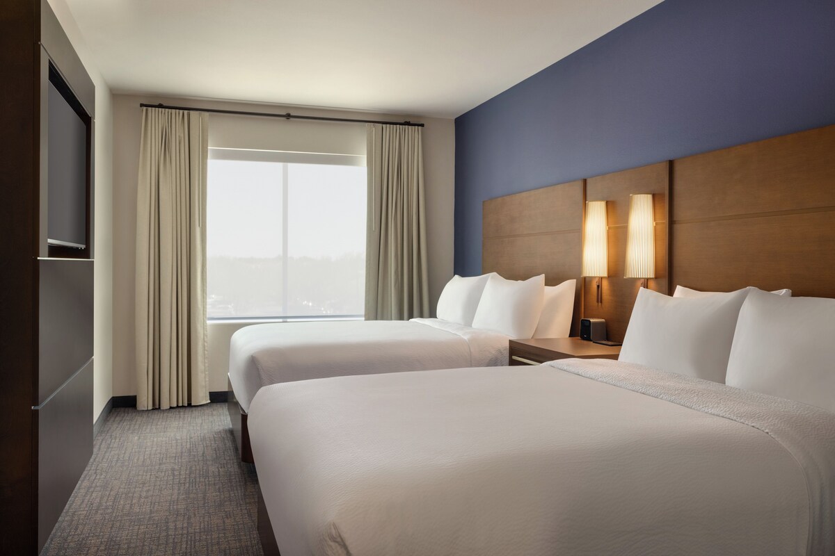 A hotel room with two beds, a large window with curtains, a flat-screen TV, and a nightstand with a lamp.