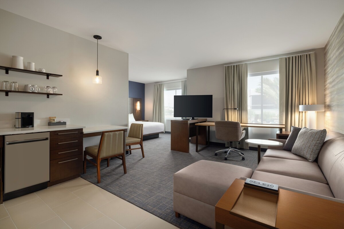 A hotel room with a kitchenette, a bed, a desk, a TV, a sofa, and a window with curtains.