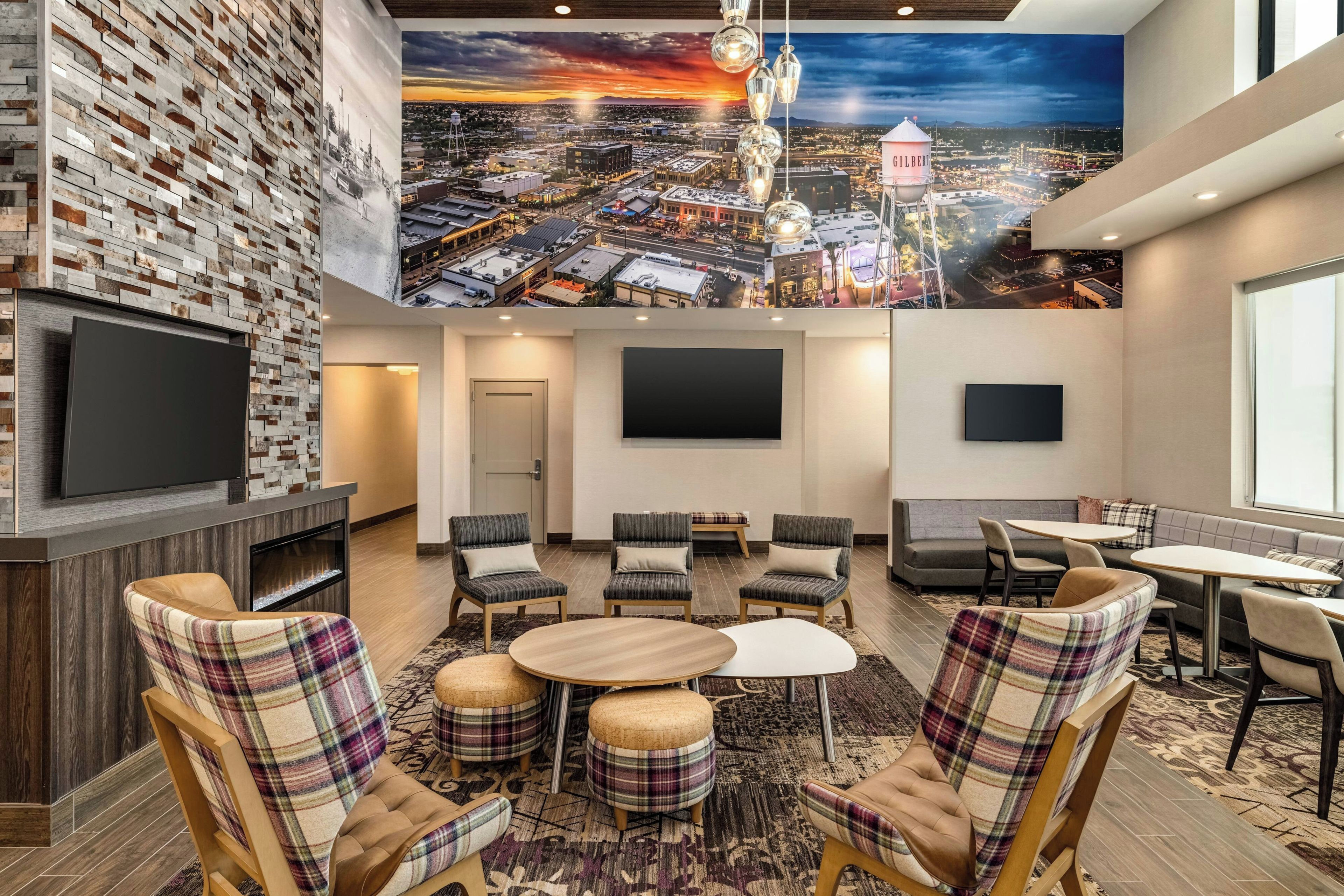 Um saguão de hotel com um mural da paisagem urbana, uma TV de tela plana, área de estar com poltronas e pufes xadrez, e uma grande janela.