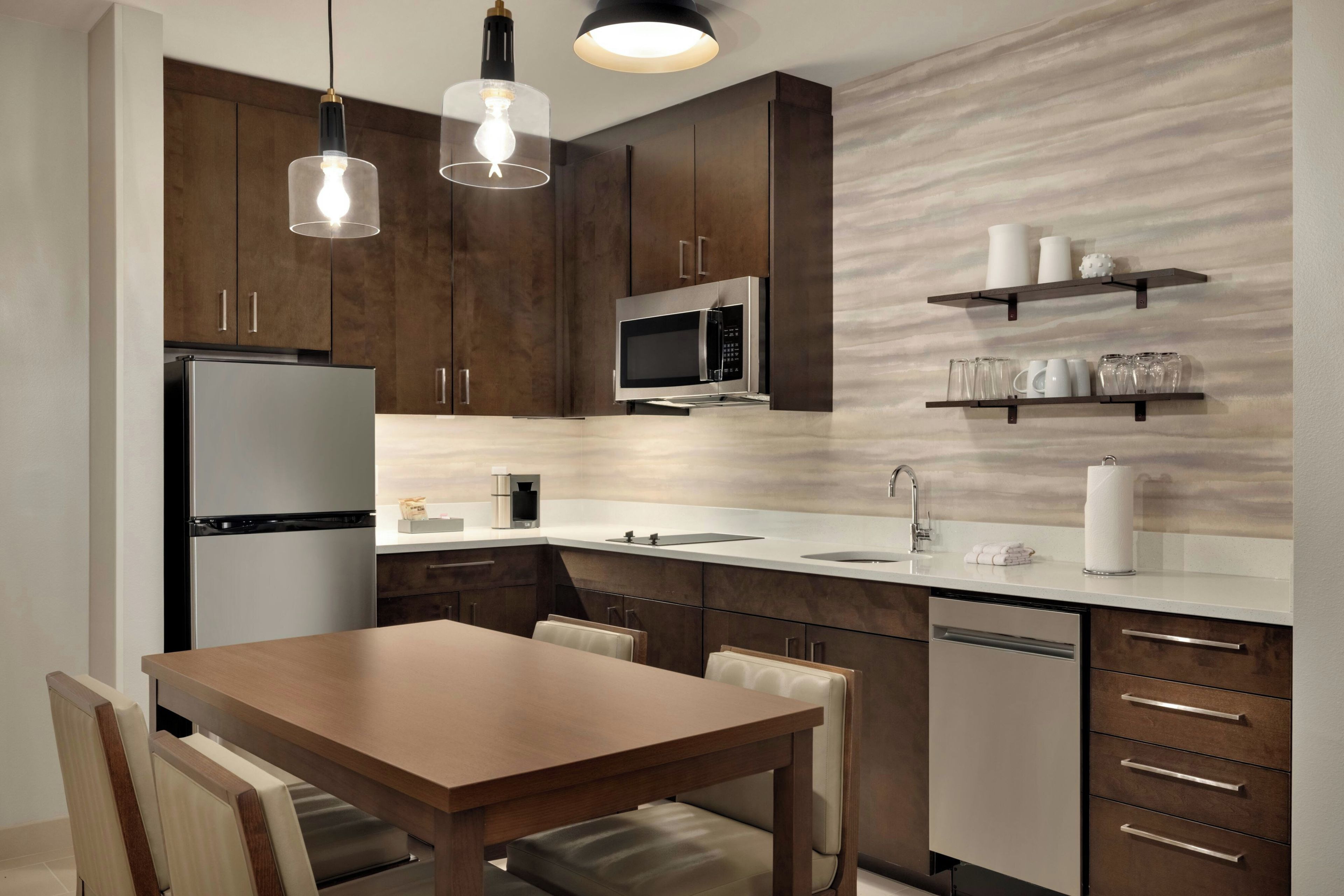 A modern kitchen with dark wooden cabinets, a stainless steel refrigerator, a microwave, and a built-in oven. There is a dining table with chairs and a countertop with a sink and a coffee machine.