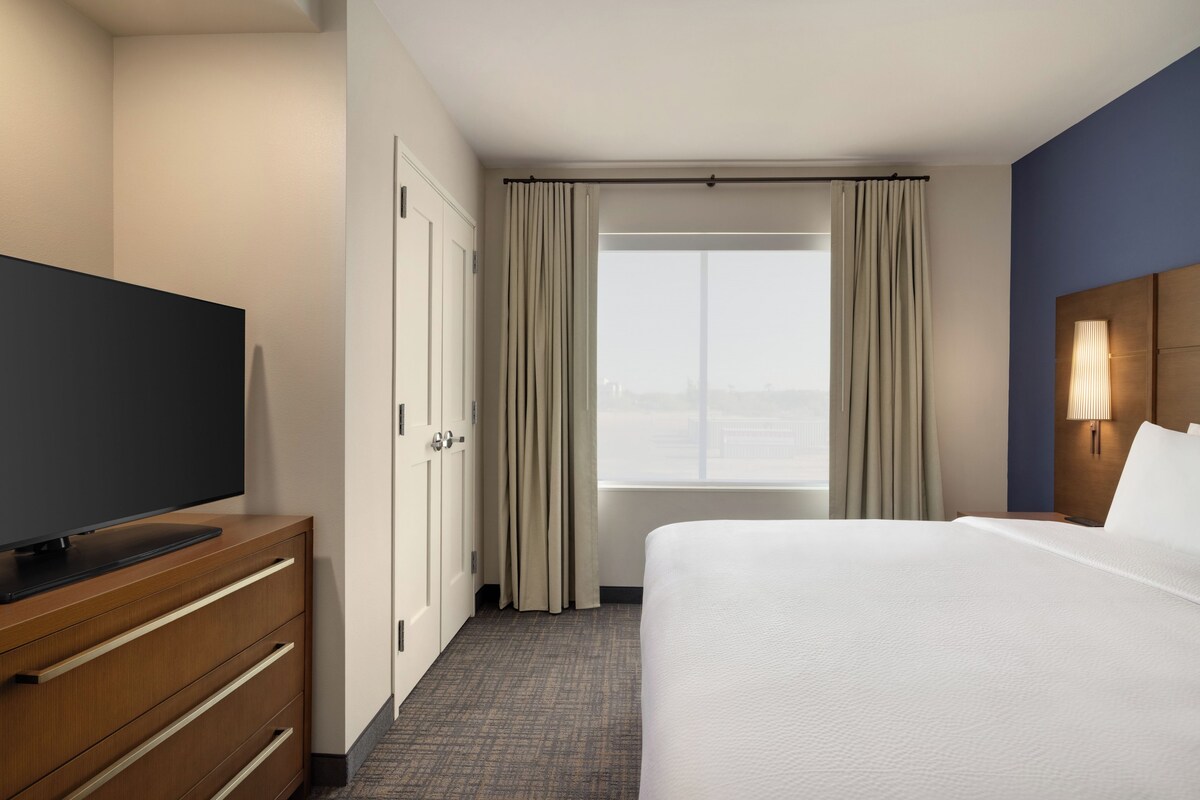 A hotel room with a bed, a television on a wooden dresser, a window with curtains, and a nightstand with a lamp.
