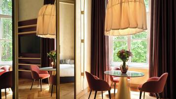 Une chambre d’hôtel avec un lit, une table ronde entourée de chaises, un vase rempli de fleurs et de grandes fenêtres avec des rideaux.