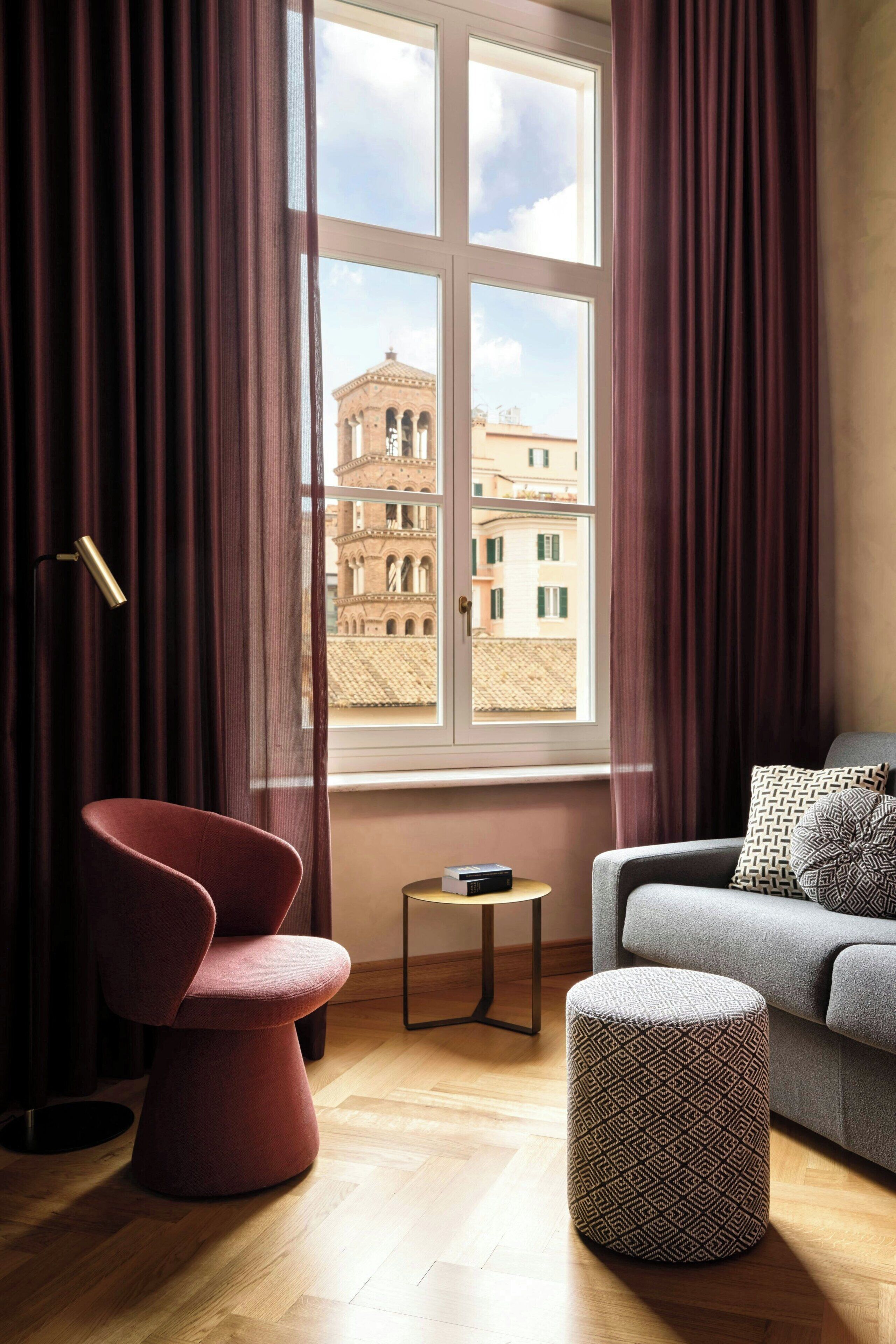 Ein Zimmer mit Blick auf ein Gebäude, ein roter Sessel, ein graues Sofa und ein gemustertes Hocker.
