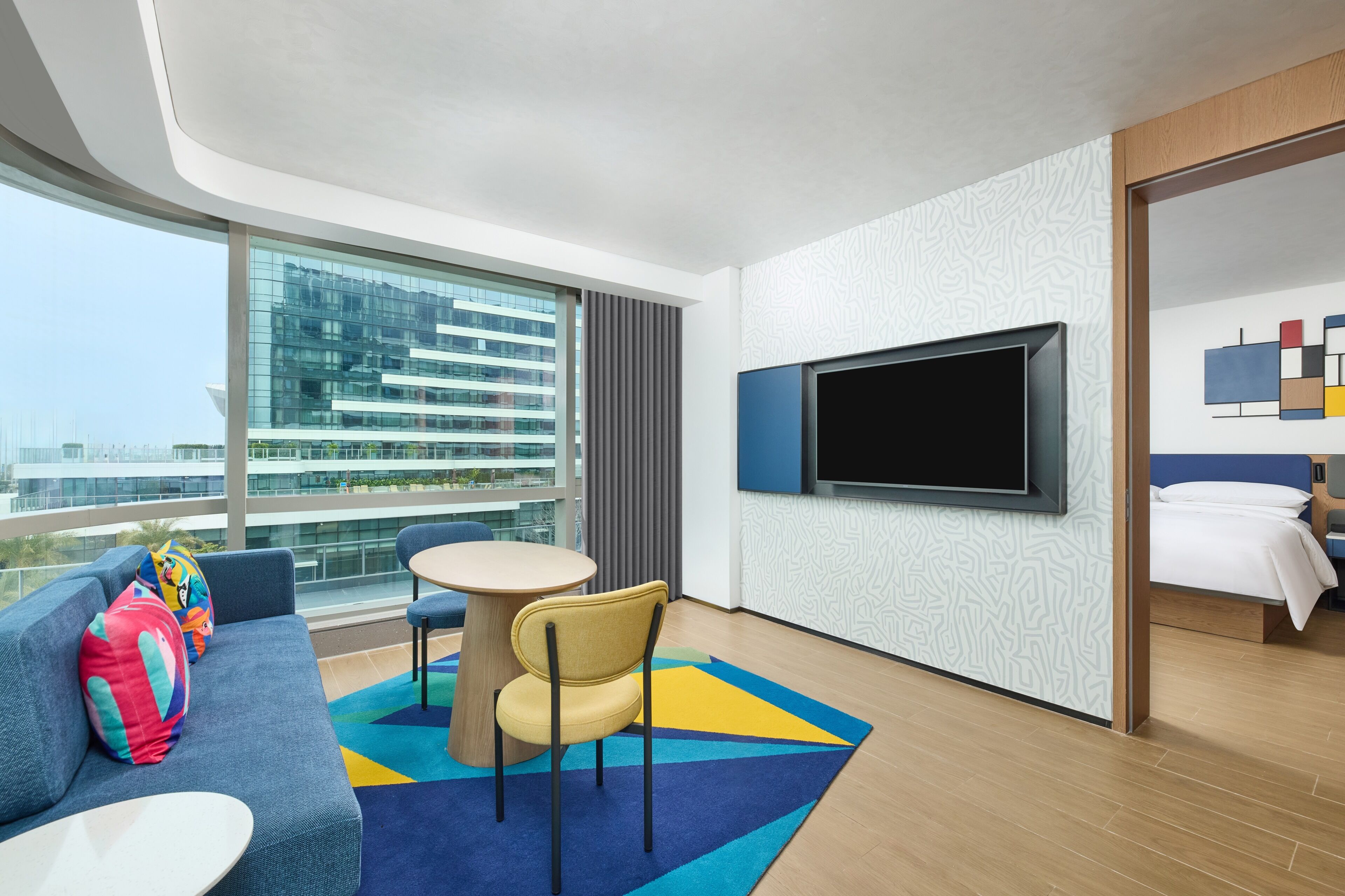 Een moderne hotelkamer met een groot raam, een flatscreen televisie, een bank met kleurrijke kussens, een ronde tafel en een bed.