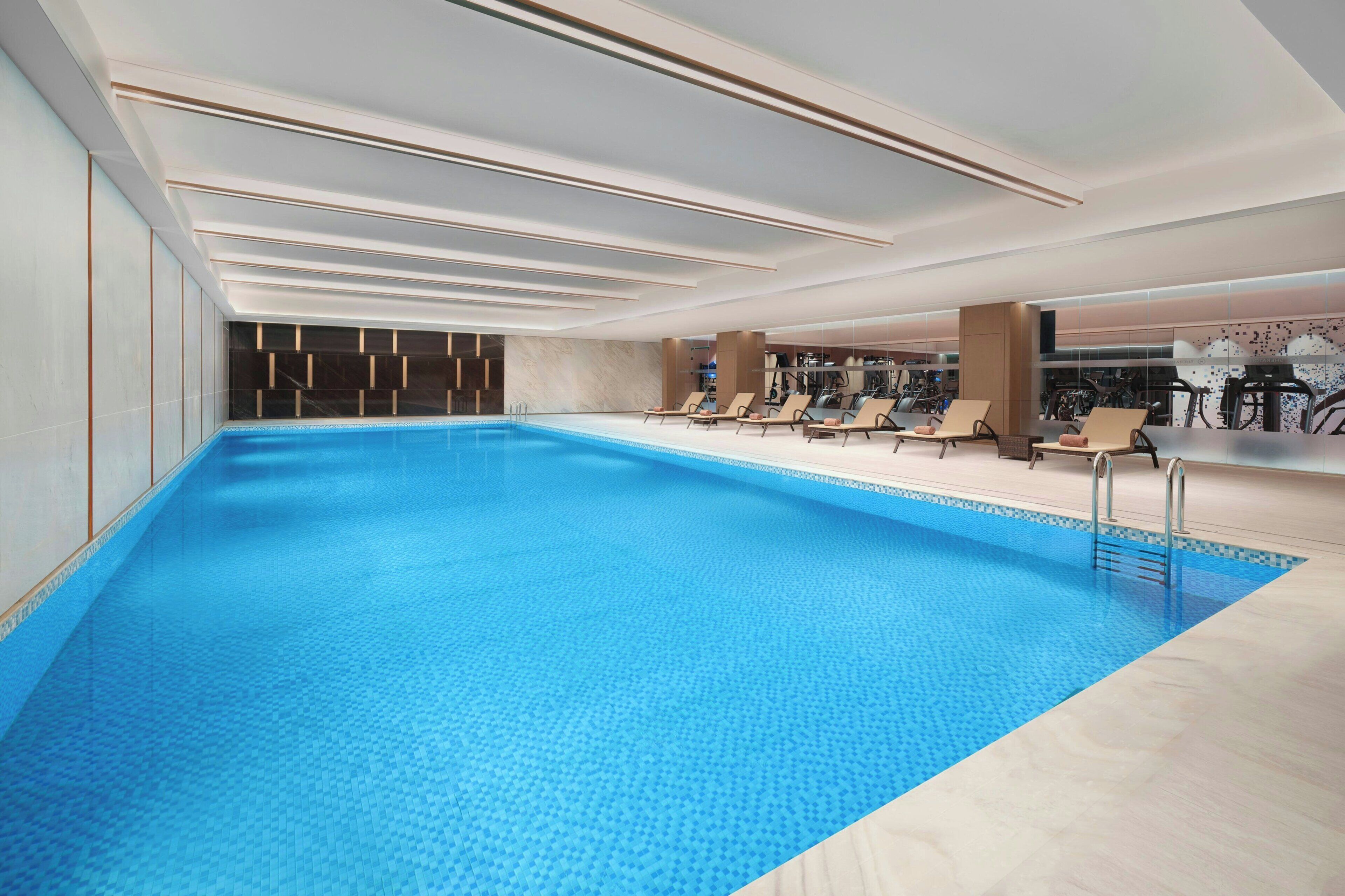 Una piscina interna moderna con superficie d'acqua blu trasparente, sedie a sdraio e attrezzature per l'esercizio fisico.