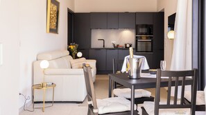 Een moderne woonkamer met een bank, een eettafel met stoelen, een keukenhoek met magnetron en oven, en een klein bijzettafeltje met lamp.