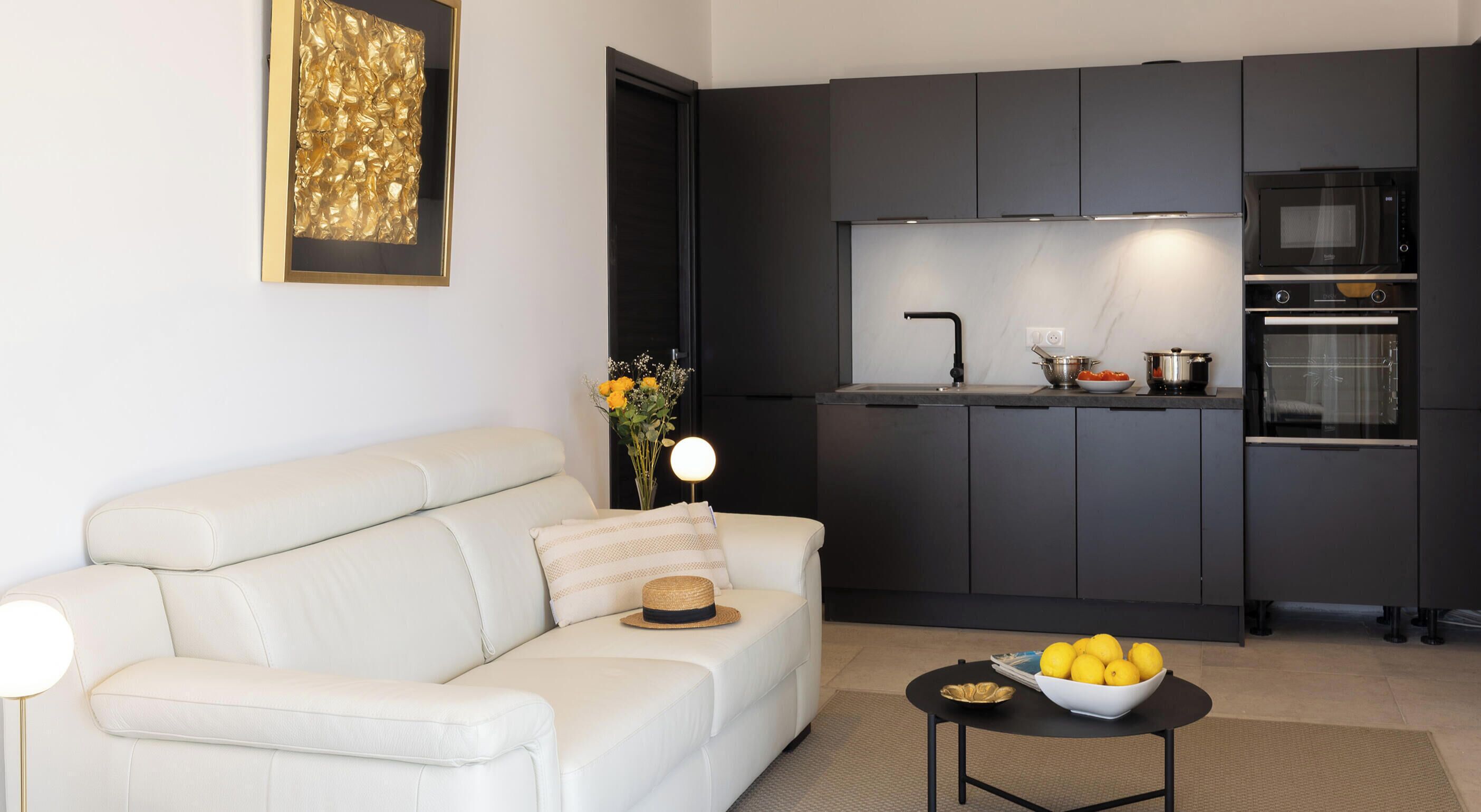 A modern living room with a sofa, a small table with a bowl of lemons, and a kitchen with dark cabinets and appliances.