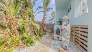 Un chemin menant à un bâtiment dont le sol est recouvert de carreaux, entouré de plantes tropicales et agrémenté d’une planche de surf décorative.