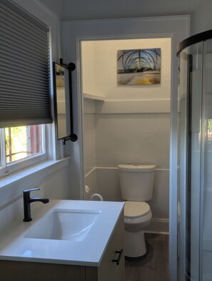 Ein Badezimmer mit einem weißen Waschbecken, einem schwarzen Wasserhahn und einer Toilette. Ein Fenster mit Jalousien und ein Bild über der Toilette sind ebenfalls vorhanden.