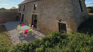 En bygning i sten med et tagtækket tag, en grusbelagt terrasse med et bord og stole, og en klar himmel med skyer.