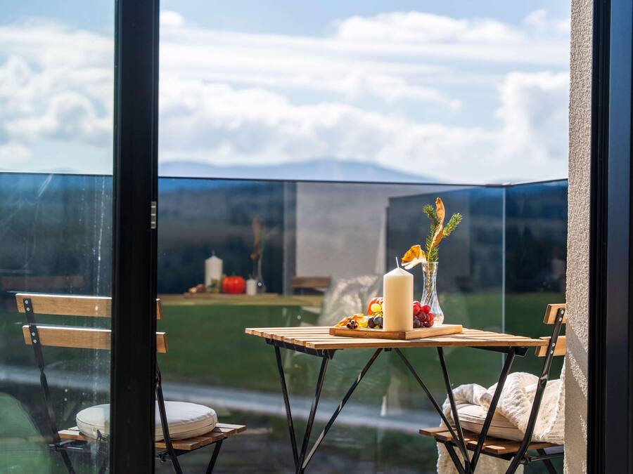 A room with a view of mountains, a table with a candle, fruits, and a vase.