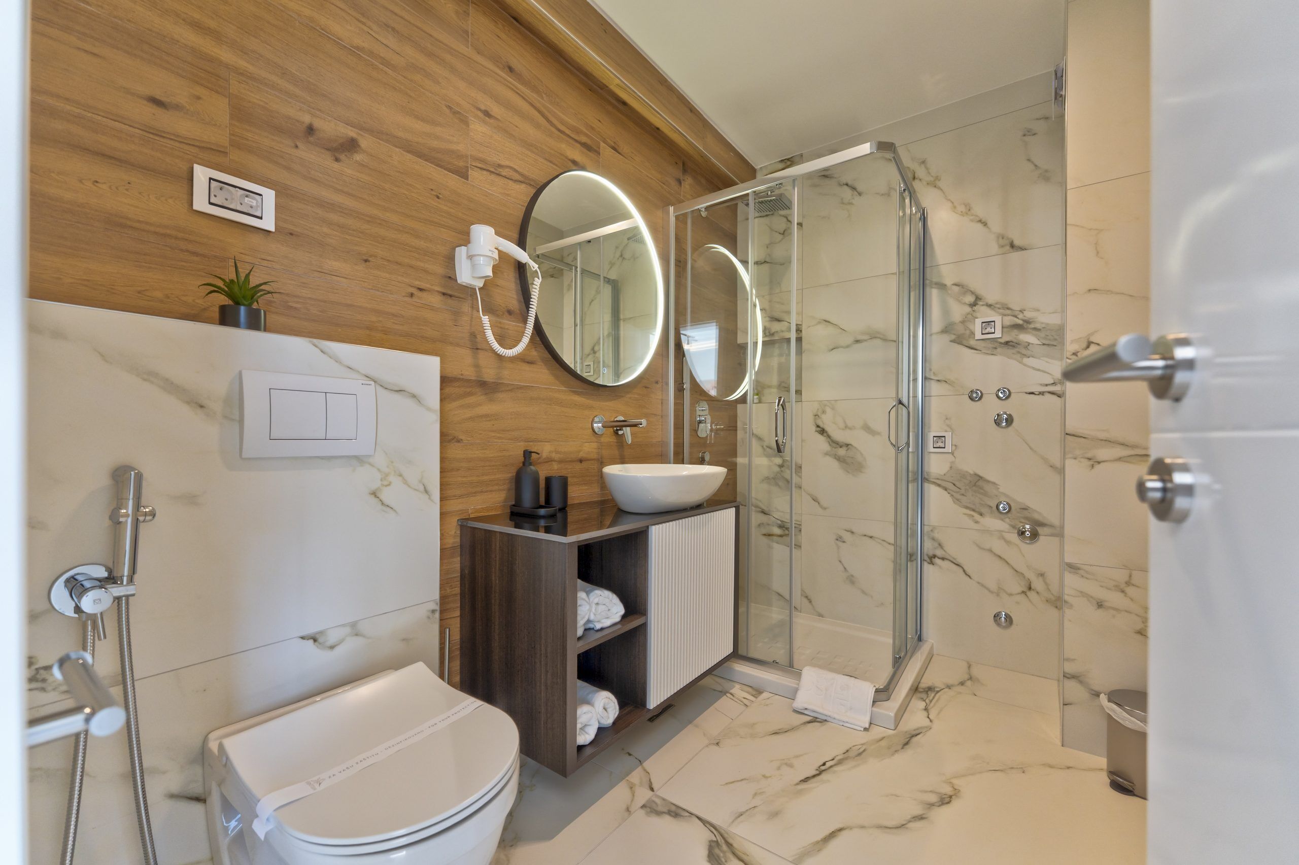 Un baño moderno con pared de madera, piso de mármol, cabina de ducha de vidrio, lavamanos blanco y un inodoro.