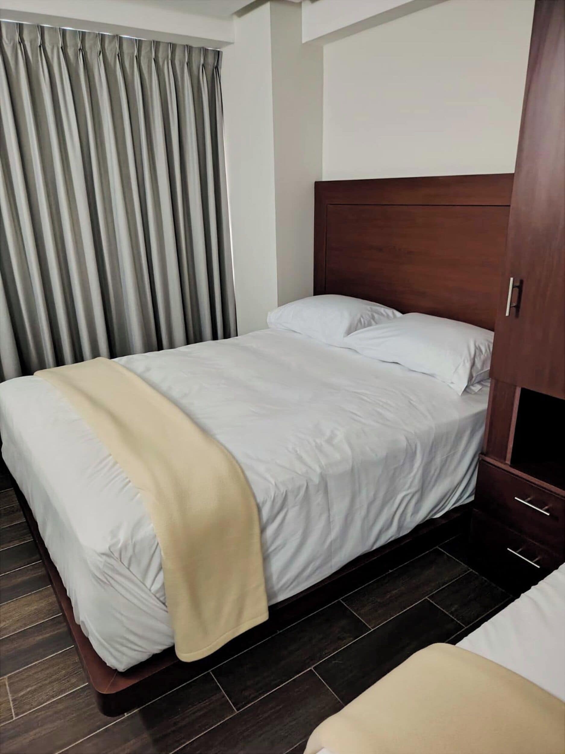 A hotel room with a bed, a wooden headboard, a beige throw blanket, and a dark wooden floor.