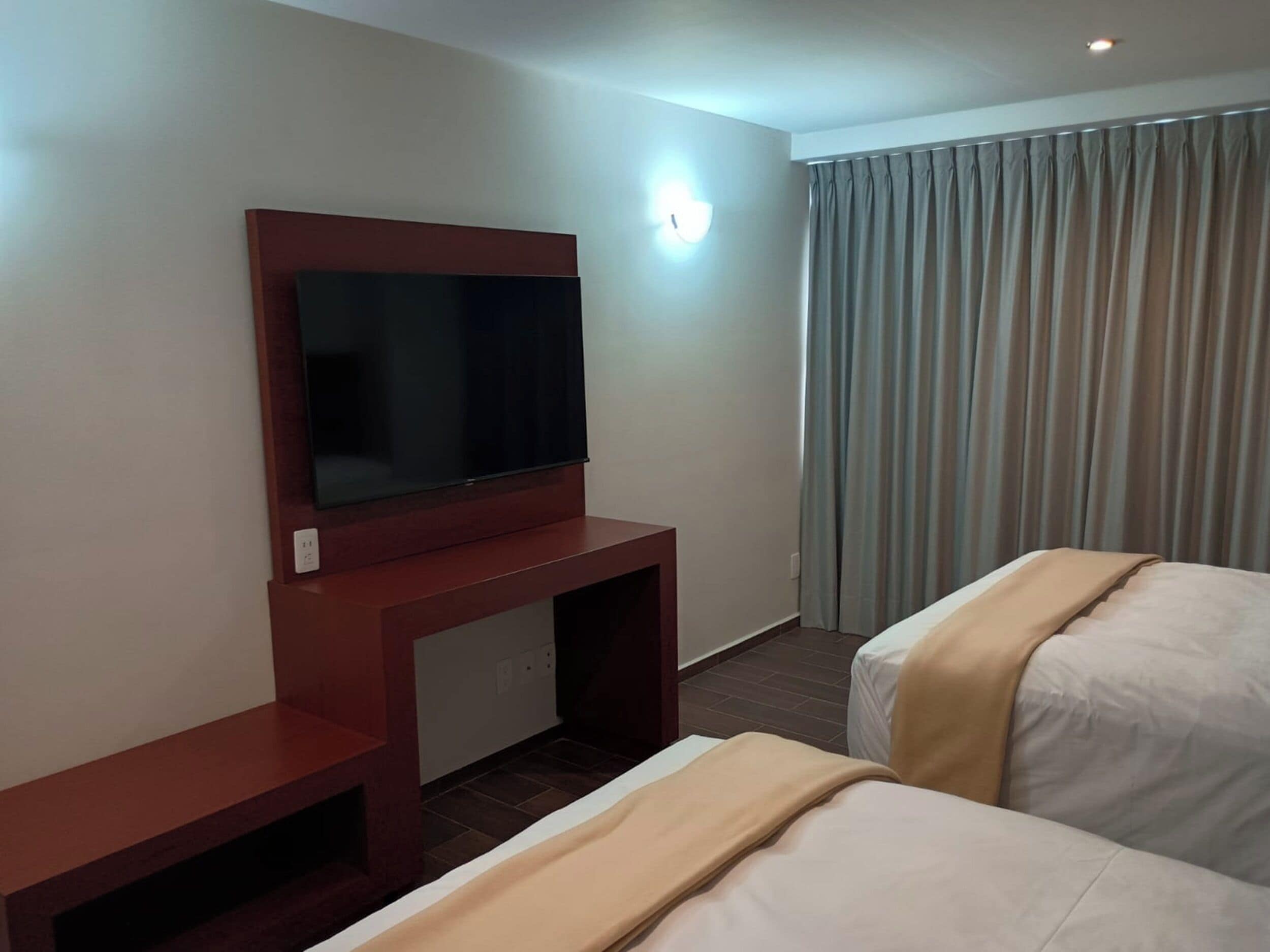 Una camera d'albergo con TV montata su un supporto in legno, un letto con coperta beige e una finestra con tende.