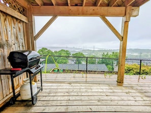 Eine Holzterrasse mit Grill und Propangasflasche, mit Blick auf eine Brücke und Grünflächen.
