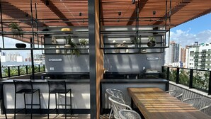 Un bar en la azotea con mesa y sillas de madera, barandilla de metal y vista panorámica de la ciudad.