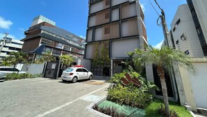 Un edificio de varios pisos con una entrada protegida por una reja, un coche aparcado y un pequeño jardín con palmeras.