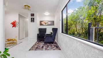 Um corredor com tapete estampado, duas cadeiras, um objeto decorativo na parede e uma janela com vista para vegetação.