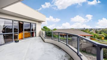 Varanda moderna com guarda-corrimão curvo, porta de vidro que dá para um espaço interior com sofá amarelo, e vista desobstruída de uma área residencial com casas e vegetação.