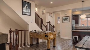 A spacious living area with a foosball table, wooden flooring, a staircase, and a kitchen area. - Girdwood Mountain Basecamp-Sleeps 7-Near Alyeska (Anchorage)