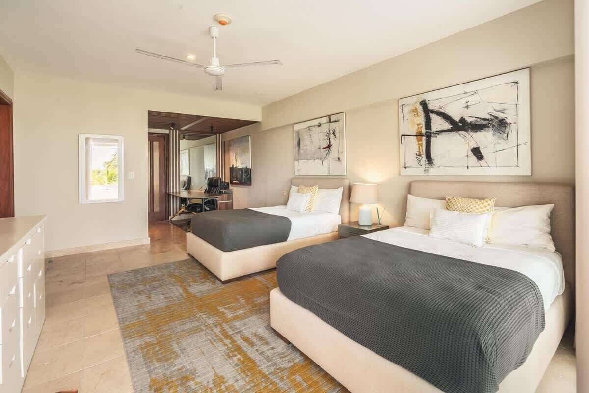 A hotel room with two beds, a ceiling fan, artwork on the walls, and a view through an open door.