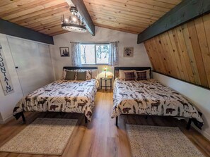 Quarto com duas camas, vigas de madeira no teto, janela com cortinas, uma mesinha com abajur e um quadro de urso na parede.
