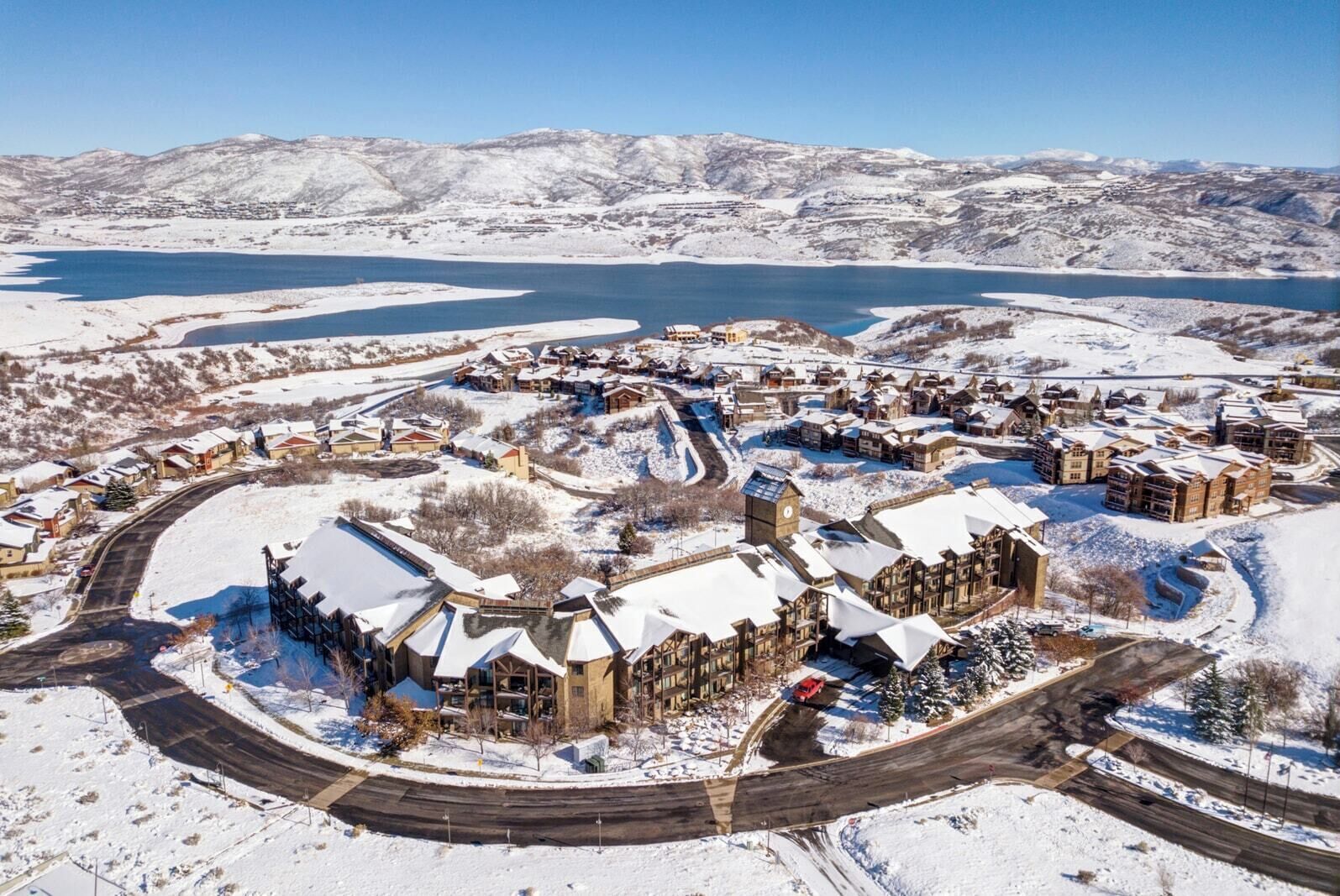 Un resort cubierto de nieve con un edificio central, un lago y montañas al fondo.