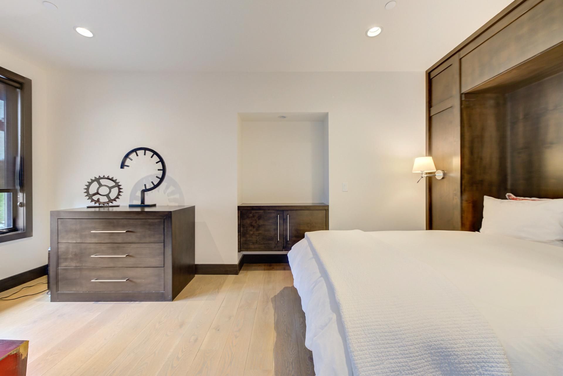 Un dormitorio con cama, cómoda, reloj de pared, mesita de noche y ventana.
