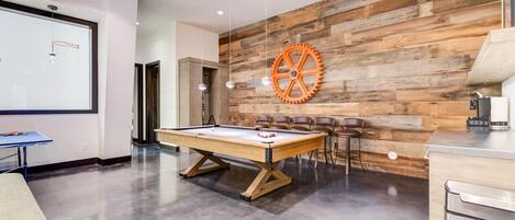 Mesa de billar con sillas, una pantalla de proyector y una cafetera en una habitación moderna con paneles de madera.