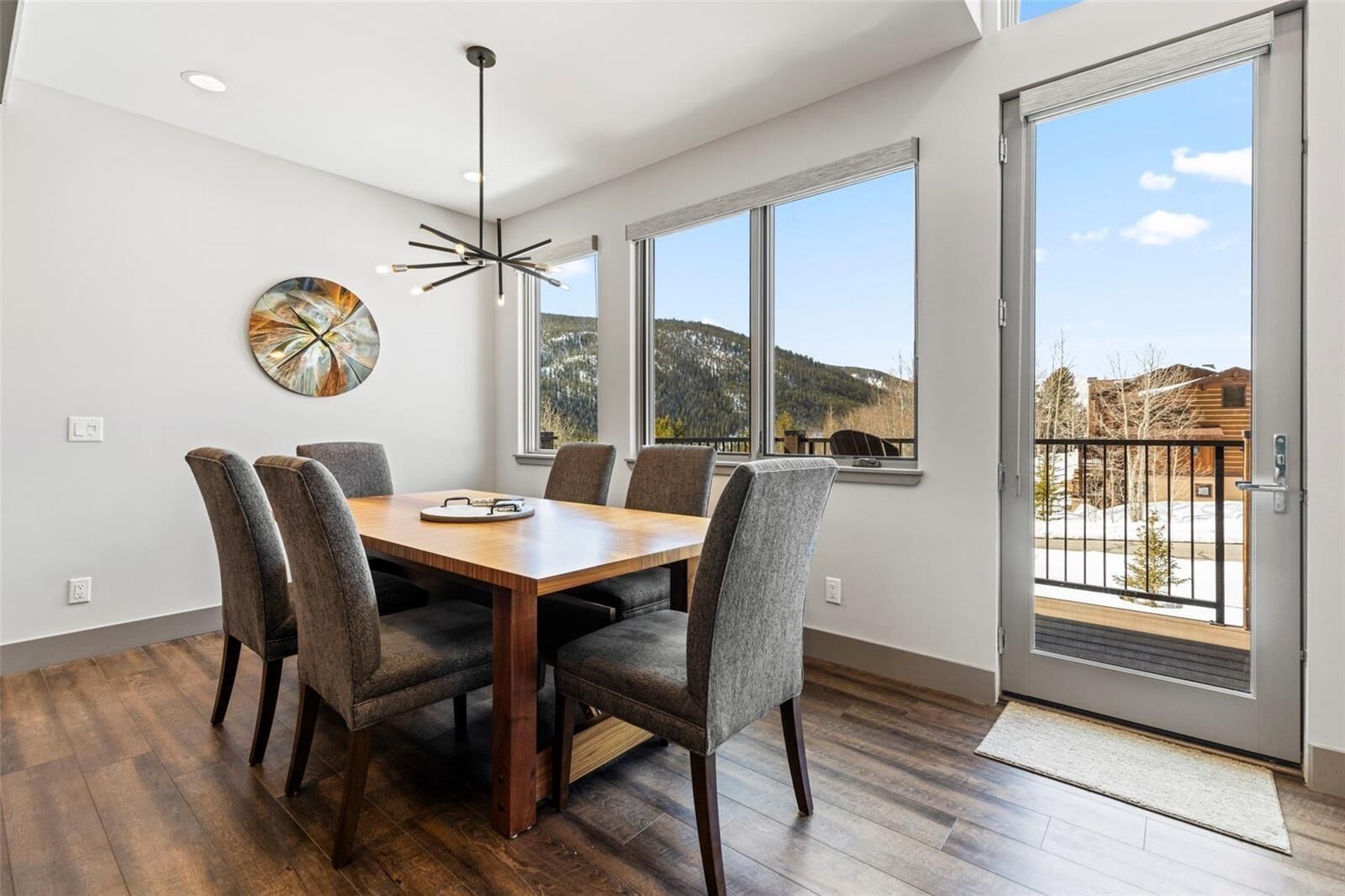 Un comedor con mesa de madera, seis sillas, una lámpara colgante moderna y un ventanal con vistas a un paisaje nevado.