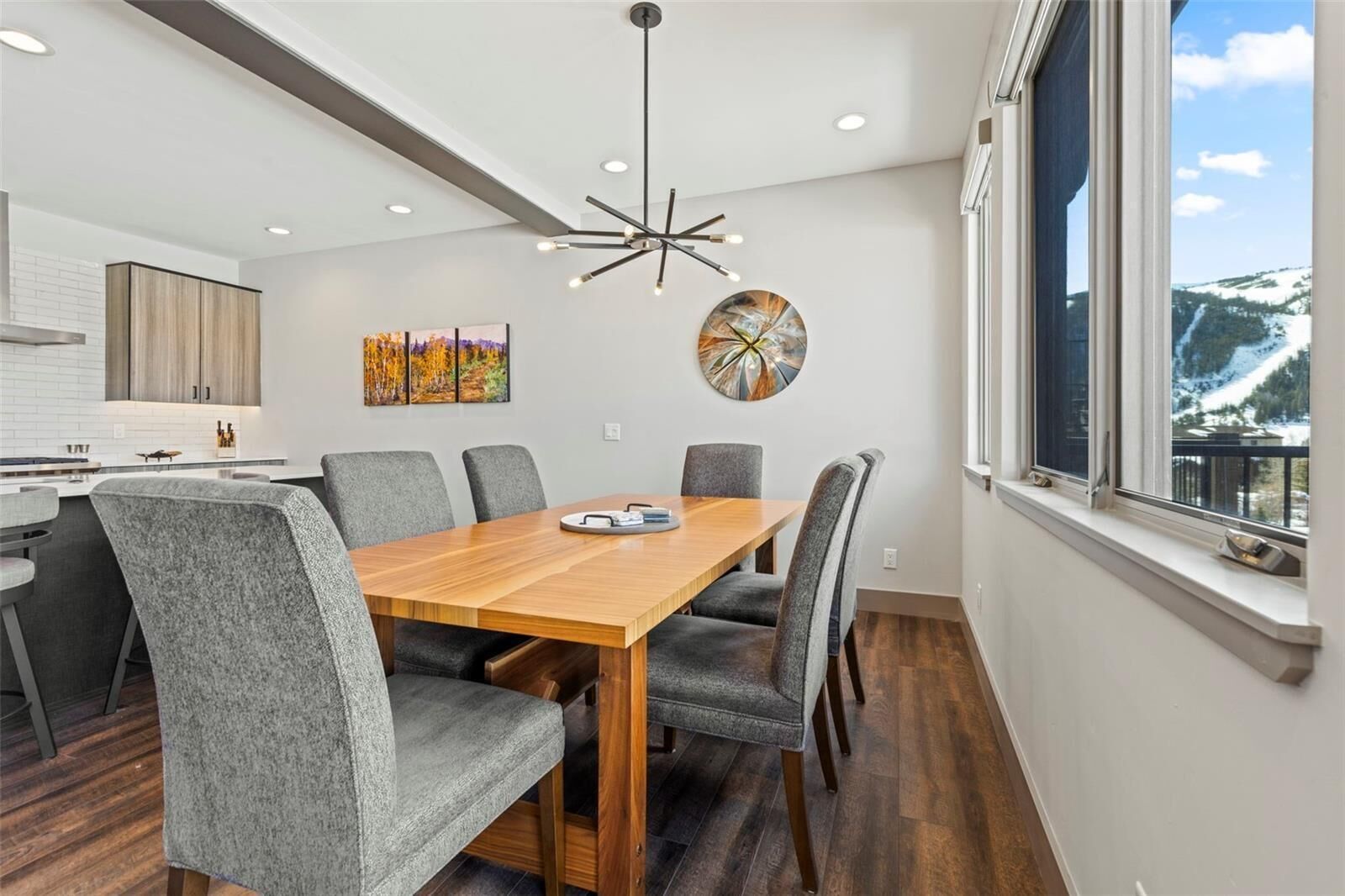 Un comedor moderno con una mesa de madera, sillas tapizadas de gris, una lámpara contemporánea y un cuadro en la pared.