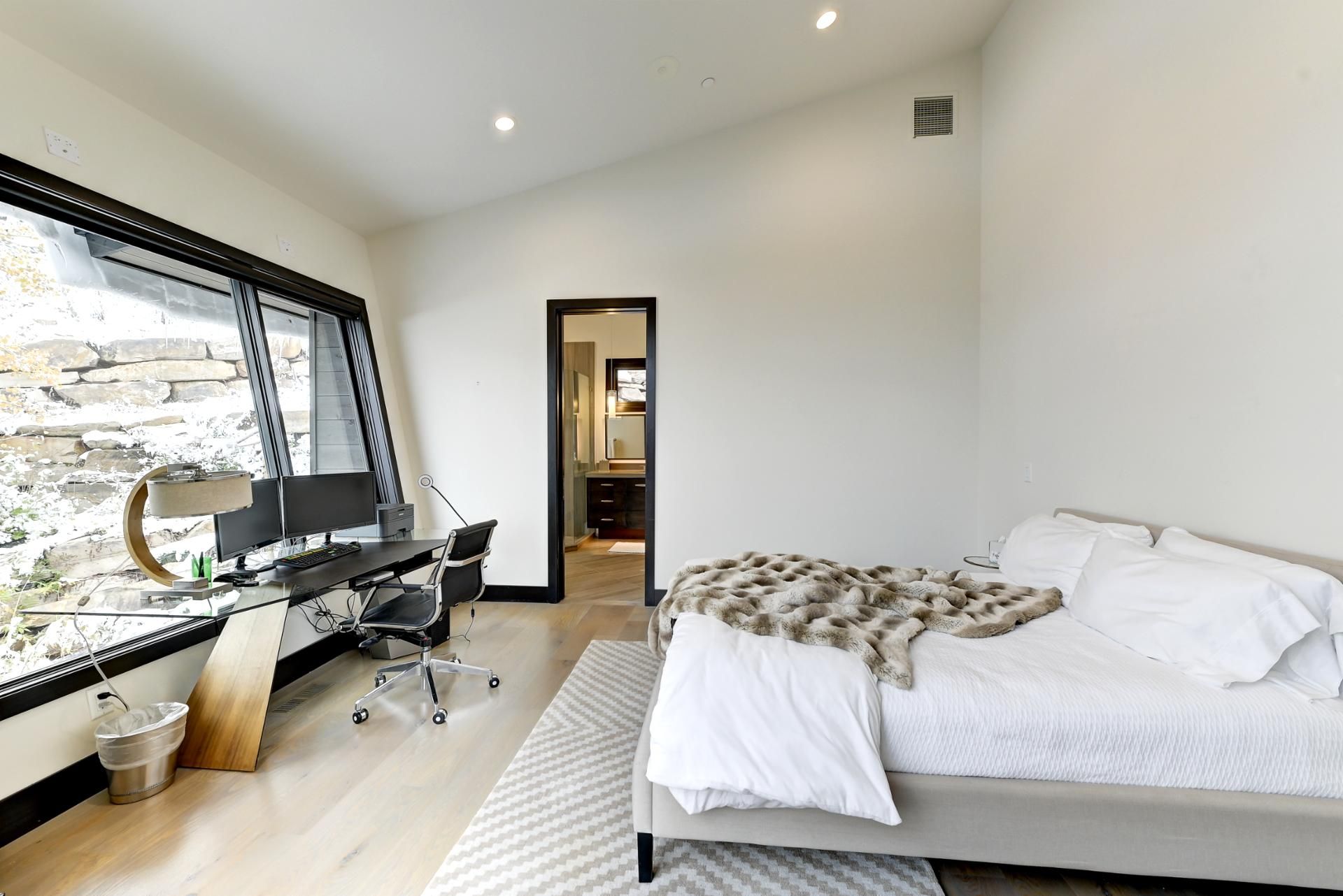 Un dormitorio moderno con una cama grande, un escritorio con computadora y vistas a un muro de piedra a través de una ventana.