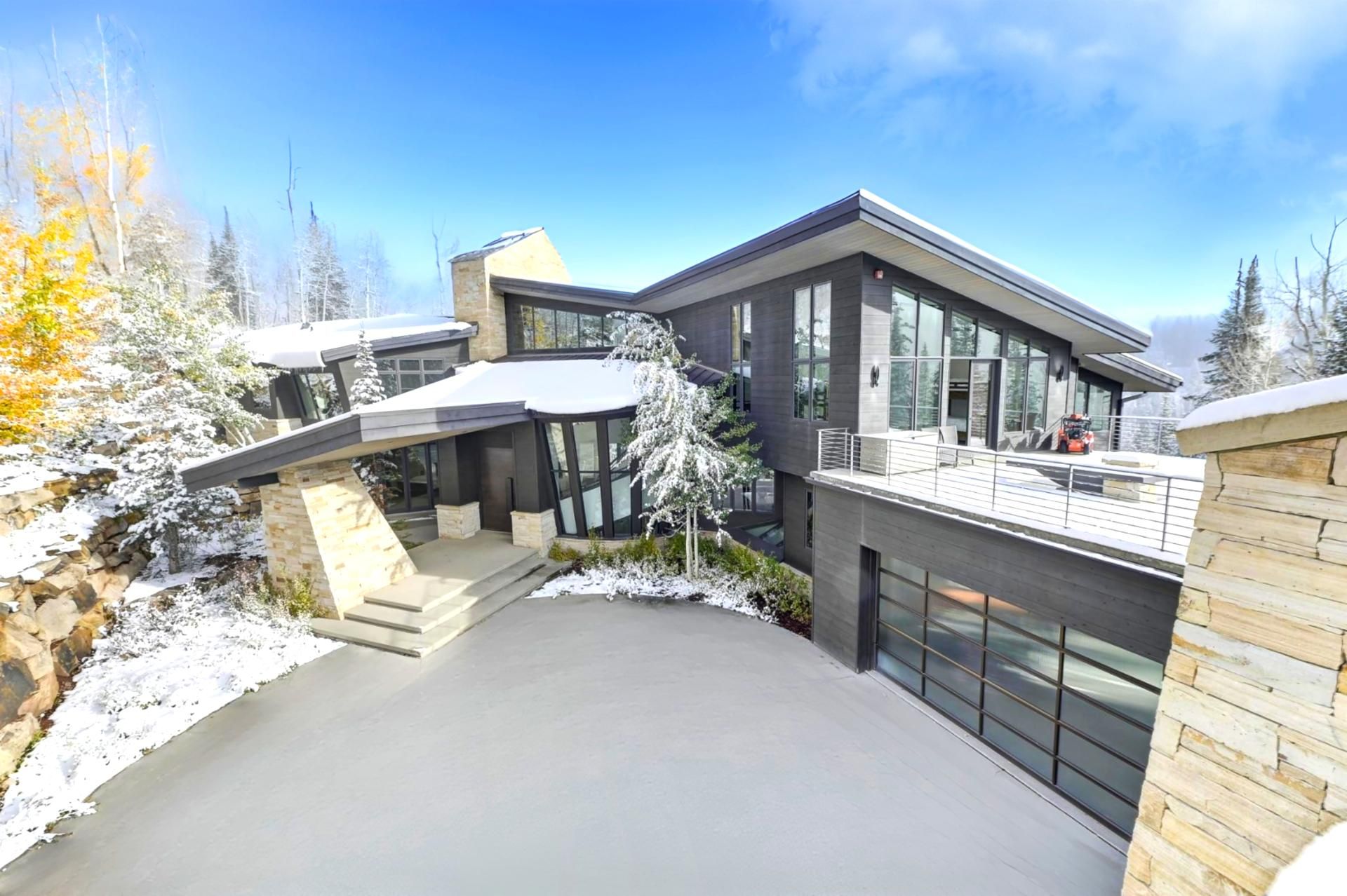 A modern multi-level house with a covered driveway, large windows, and a view of snow-covered trees.