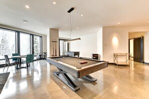 A modern indoor pool table with a wooden top and metal frame, a dining area with chairs and a table, and large windows offering a view of trees. - KBM Resorts| Walk to Slopes, Sports Court, Private Home Theater, Sleeps 17, Outdoor Hot Tub PCO-184M (Park City)