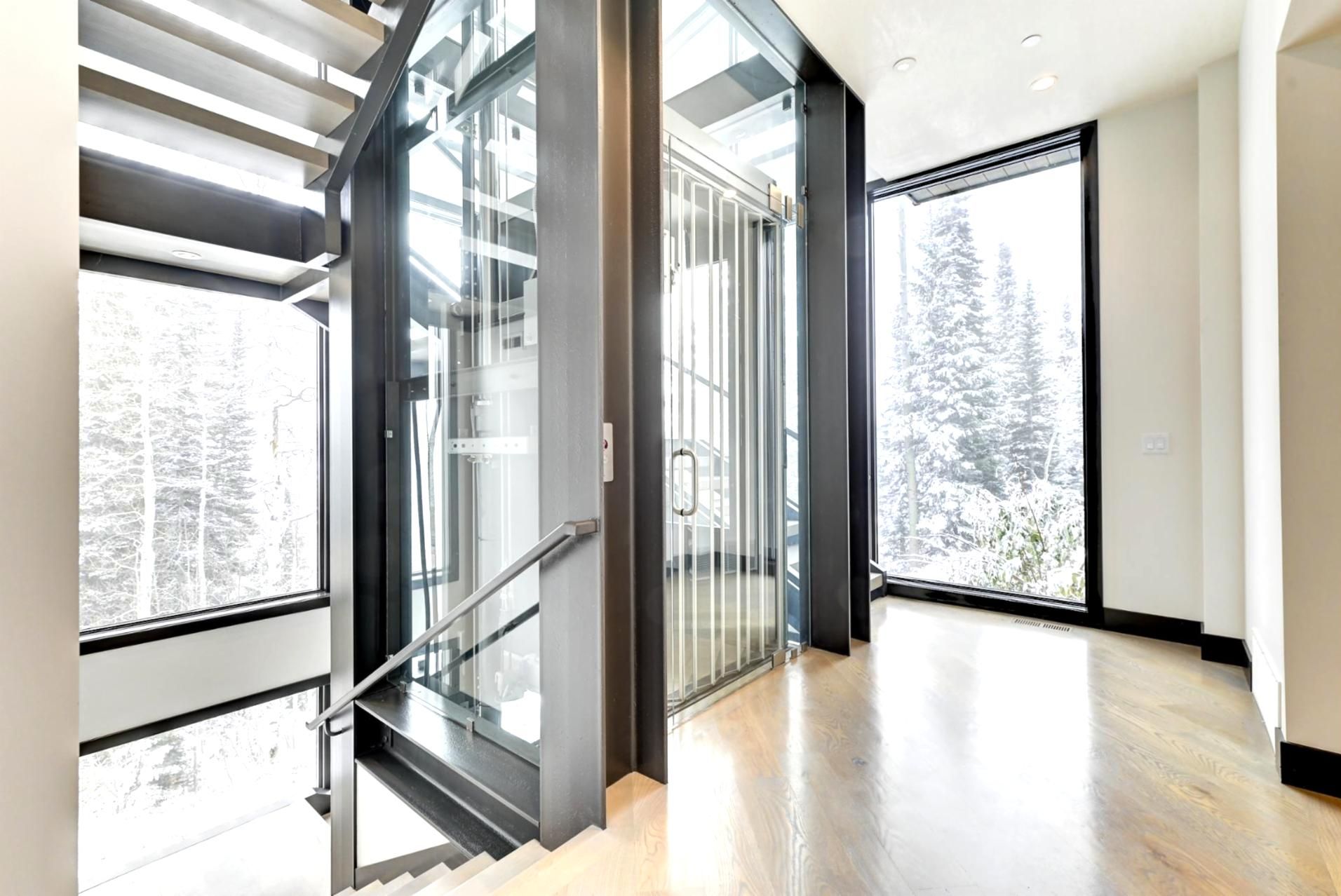Un interior moderno con una escalera que lleva a un balcón, amplias ventanas que ofrecen vistas a árboles, y un piso de madera pulida.