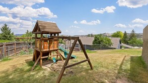 Estructura de madera para juegos con tobogán y columpio en un jardín cubierto de césped.
