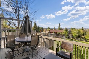 A deck with outdoor furniture, a table, and chairs, surrounded by a wooden railing with a view of houses and trees in the background.