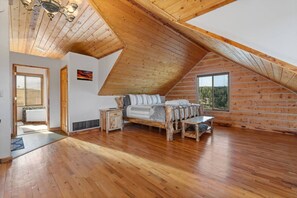 Een slaapkamer met een schuine kap, houten vloer, een bed met houten hoofdeinde, een nachtkastje en een raam met uitzicht op bomen.