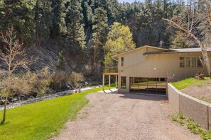 A two-story wooden house with a gravel driveway and a covered outdoor area, surrounded by a forested hillside. - Creekside Retreat | Fireplace + Gameroom (Rapid City)